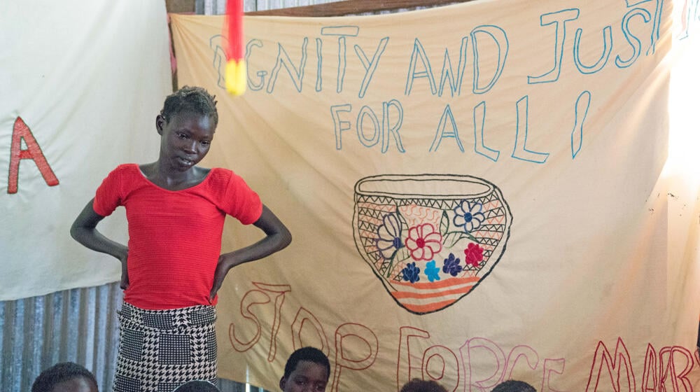 A young girl stands in front of a banner that reads "Dignity and Justice for All" and "Stop Forced Marriage,